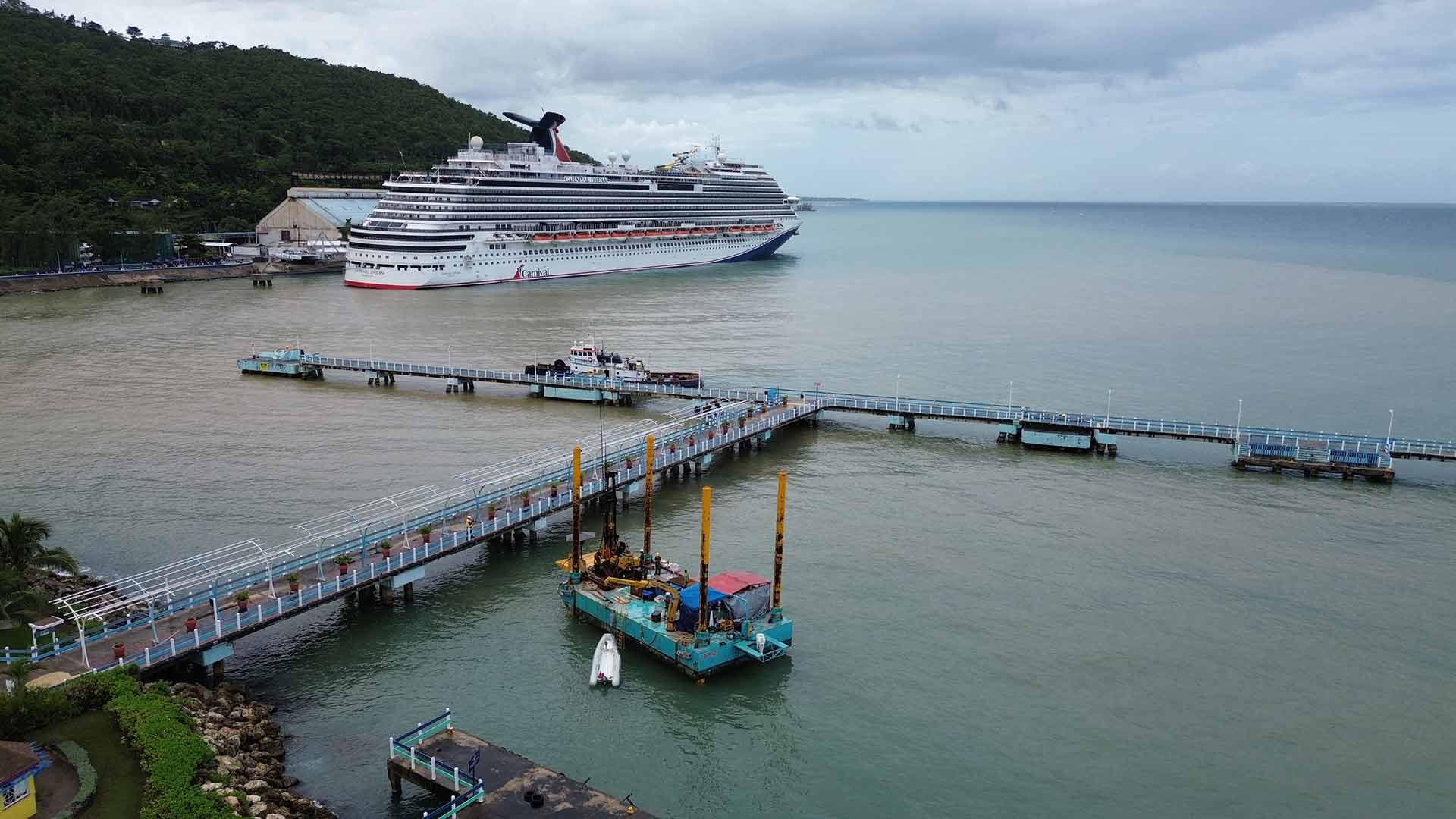 The Ocho Rios cruise ship terminal as seen from the above the shore. Looking out on the water, a Carnival cruise ship can be seen anchored a short distance away. In the foreground the Harbourside geotechnical barge is anchored near the pier.